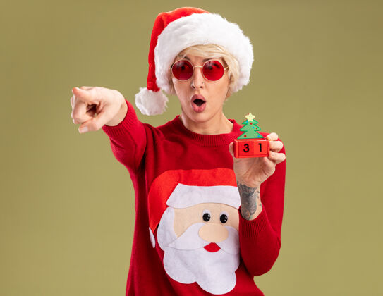 帽子惊讶的年轻金发女人戴着圣诞帽和圣诞老人的圣诞毛衣戴着眼镜拿着圣诞树玩具和日期看 并指着一边孤立的橄榄绿墙上的复制空间指向壁板圣诞老人