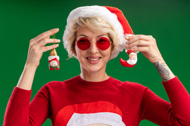 帽子微笑的金发女郎戴着圣诞帽和圣诞老人的圣诞毛衣 戴着眼镜 看起来像是雪人和圣诞老人的圣诞饰品孤立在绿色的墙上圣诞老人雪人穿