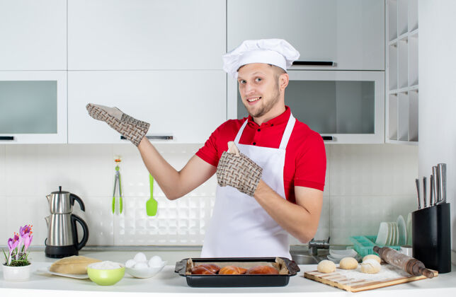 帽子俯视图：自信的男厨师戴着支架站在桌子后面 上面放着糕点鸡蛋磨碎器 在白色厨房的右侧展示着一些东西厨房磨碎机东西