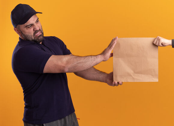 衣服一个穿着制服 戴着帽子的中年送货员把纸食品包装交给隔离在黄色墙上的客户给予姿势立场