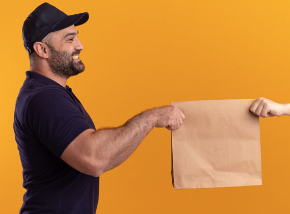 人微笑着站在剖面图中年送货员在制服和帽子给纸食品包装客户隔离在黄色的墙上微笑包装表达