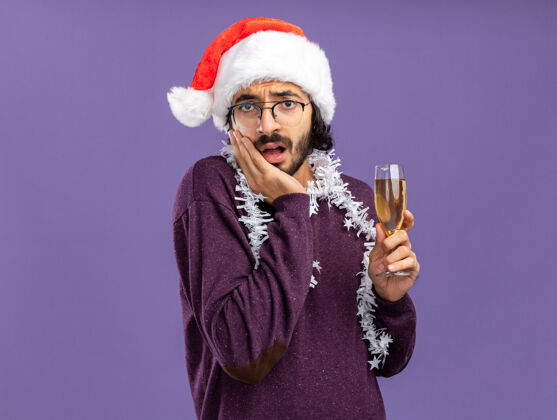 帅气年轻帅哥戴着圣诞帽 脖子上戴着花环 手里拿着一杯香槟 手放在脸上 被隔离在蓝色的墙上杯子圣诞节抱着