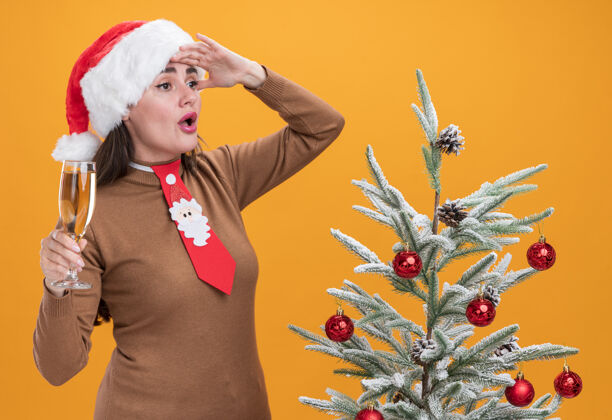 立场惊讶的年轻漂亮女孩戴着圣诞帽打着领带站在圣诞树旁拿着一杯香槟望着远方 手被隔离在橙色的墙上举行杯子年轻