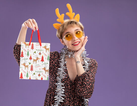 圣诞节微笑着的年轻漂亮女孩戴着驯鹿鹿角头带 脖子上戴着金箔花环戴着眼镜拿着圣诞礼品袋手放在脸上看着紫色墙上孤立的一面 还有复制空间驯鹿脖子鹿角