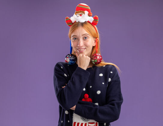 穿微笑的年轻漂亮女孩穿着圣诞毛衣 戴着圣诞发箍 耳朵上挂着圣诞球 手放在下巴下面 隔离在紫色的墙上圣诞节年轻毛衣
