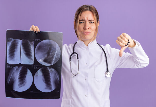 听诊器年轻的女医生穿着医用长袍 听诊器拿着x光片 拇指朝下 被隔离在紫色的墙上向下衣服人