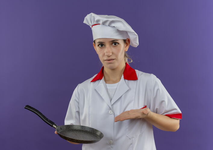 指着令人印象深刻的年轻漂亮厨师在厨师制服举行 并用手指着煎锅隔离在紫色的墙壁厨师漂亮手