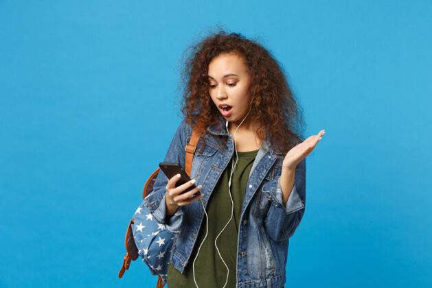 女性年轻的非洲裔美国少女 穿着牛仔服 背着耳机 隔离在蓝色的墙上电话举行音乐