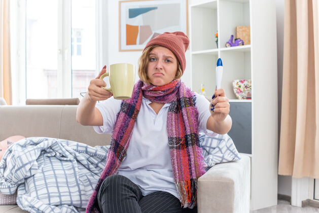周围不健康的年轻女子戴着暖和的帽子 脖子上围着围巾 手里拿着一杯茶和温度计 坐在明亮的客厅里的沙发上 看起来很担心温暖担心温度计