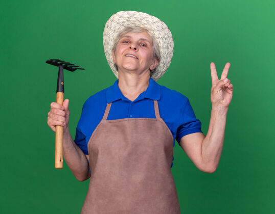 花园自信的上了年纪的女园丁戴着园艺帽拿着耙子 打着胜利的手势女性穿老人