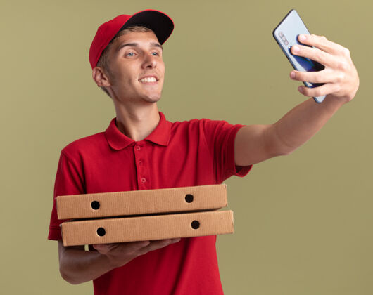 自拍微笑的年轻金发送货男孩拿着比萨饼盒 看着手机自拍金发盒子披萨