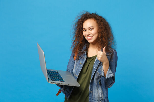 持有年轻的非洲裔美国女孩十几岁的学生在牛仔服装 背包工作的pc隔离在蓝色的墙壁上青少年打字电脑