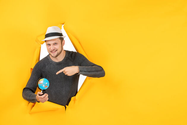 男孩一个微笑的男人 戴着帽子 手里拿着一个小地球仪 指着一面撕破的黄色墙上的一个小地球仪帽子地球仪孩子