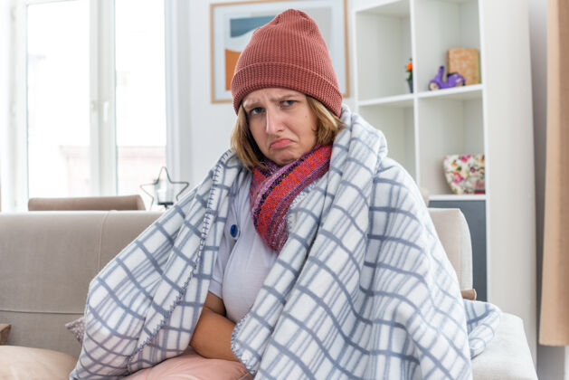痛苦不健康的年轻女子 戴着暖和的帽子 裹着毯子 看起来不舒服 生病 感冒和流感 体温计发烧 坐在客厅的沙发上 看起来很担心不适毯子流感