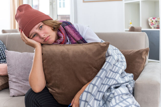 脖子不健康的年轻女子戴着帽子 脖子上围着暖和的围巾 感觉不舒服 生病 抱着枕头 患感冒和流感 表情悲伤地坐在客厅的沙发上悲伤不适帽子