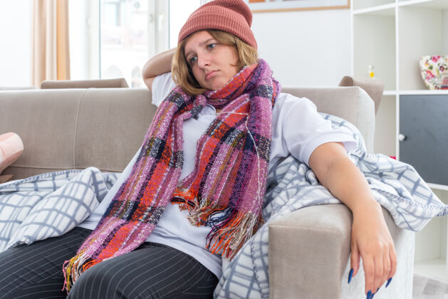 流感不健康的年轻女子戴着暖和的帽子 脖子上围着围巾 感觉不舒服 生病 患感冒和流感 看起来很担心 坐在客厅里的沙发上脖子感觉生活