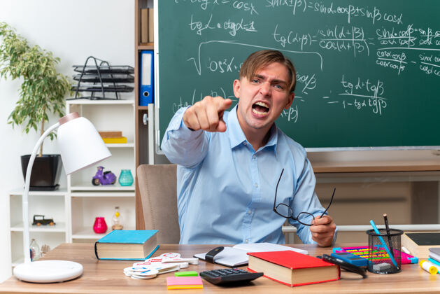 表情愤怒的年轻男老师戴着眼镜 用食指指着 带着咄咄逼人的表情大喊大叫 坐在课桌旁 教室的黑板前放着书和笔记穿喊目录