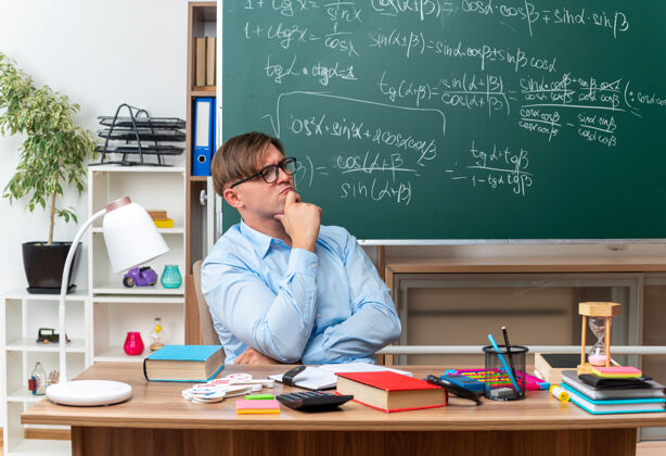 一旁年轻的男老师戴着眼镜 手放在下巴上 边看边沉思 坐在课桌旁 教室里的黑板前放着书和笔记表情学校教室
