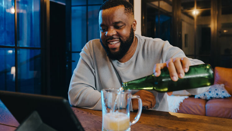 平板电脑中年非洲裔美国男性在家中客厅通过视频通话 喝啤酒 享受快乐之夜派对活动在线庆祝活动大笑男人黑人