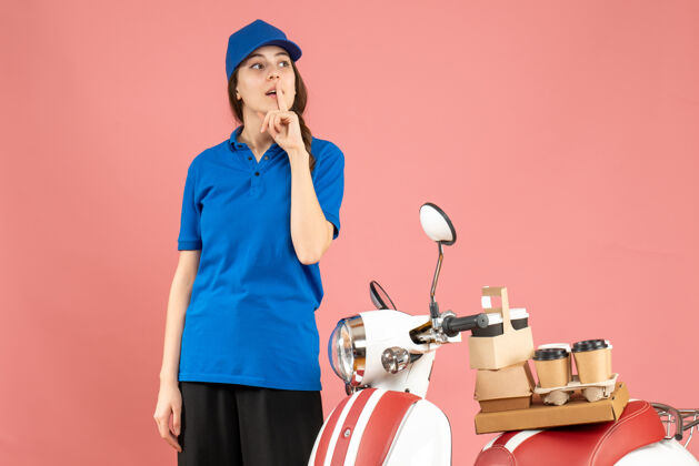 咖啡俯视图：快递员女士站在摩托车旁边 车上放着咖啡和小蛋糕 在柔和的桃色背景下做着沉默的手势人沉默桃子