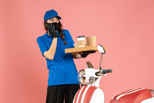 咖啡俯视图：害怕快递员的女孩戴着医用口罩手套站在摩托车旁拿着咖啡小蛋糕 背景是柔和的桃色男医疗面具