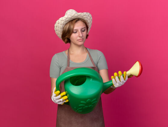 帽子困惑的年轻斯拉夫女园丁戴着园艺帽和手套拿着和看着浇水罐花园手套粉红色