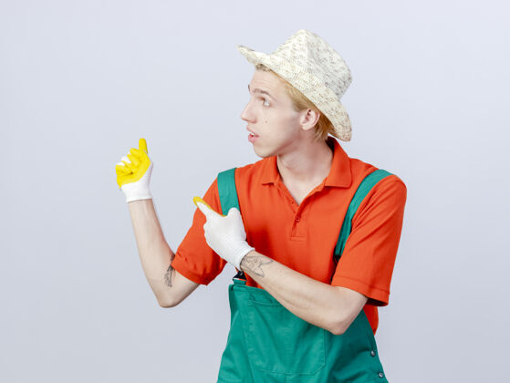 帽子年轻的园丁 穿着连体衣 戴着橡胶手套 食指指向一边立场橡胶目录