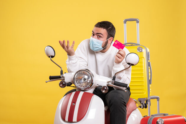 医疗旅行概念的正面图 戴着医用面罩的好奇的年轻人坐在摩托车上 黄色手提箱在上面 手里拿着银行卡男抱着手提箱