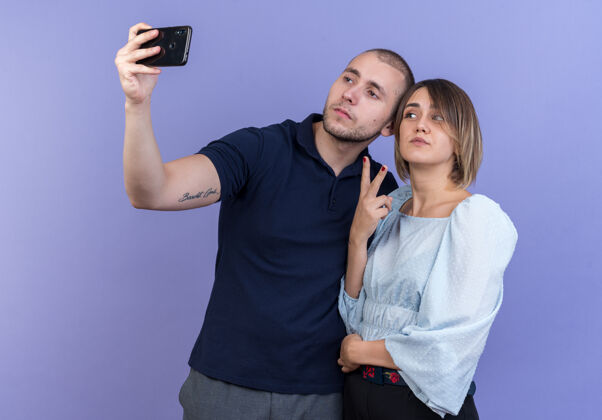 手机年轻漂亮的情侣男女快乐而积极地用智能手机在蓝色的墙上展示v型标志一起自拍男人Vsign年轻