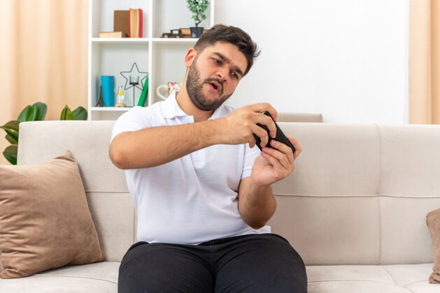 男人一个穿着休闲服的年轻人坐在客厅里的沙发上 用智能手机玩着游戏 情绪激动 兴致勃勃游戏衣服玩
