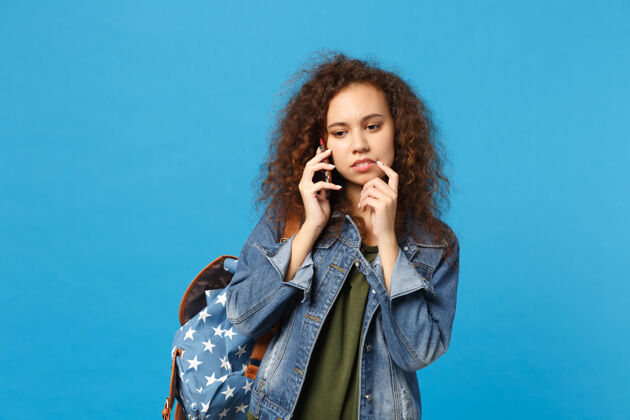 包年轻的非洲裔美国女孩十几岁的学生在牛仔服装 背包举行电话隔离在蓝色的墙壁上电话女人沮丧