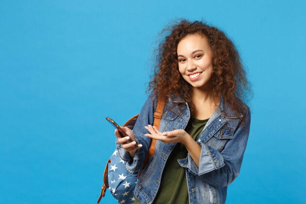 短信年轻的非洲裔美国女孩十几岁的学生在牛仔服装 背包举行电话隔离在蓝色的墙壁上青少年站设备