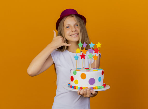 橘子戴节日帽的漂亮小女孩拿着生日蛋糕微笑着打电话给我的手势 生日派对的概念蛋糕女孩手势