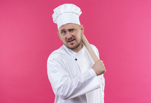 年轻自信的年轻帅哥厨师穿着厨师制服拿着擀面杖孤立地站在粉红色的墙上制服滚持枪