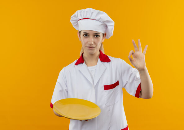 制服自信的年轻漂亮厨师 身着厨师制服 拿着盘子 在橙色的墙上贴着“一切正常”的牌子好的拿着自信
