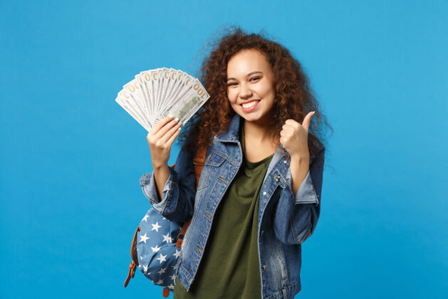 银行年轻的非洲裔美国女孩十几岁的学生在牛仔服装 背包举行现金钱隔离在蓝色的墙壁上金融包成功