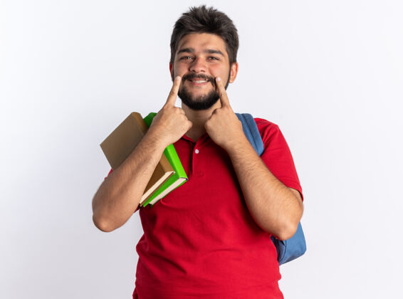 站年轻的留着胡子的学生 穿着红色马球衫 背着背包 拿着笔记本 手指着他站在白墙上的笑容微笑马球背包