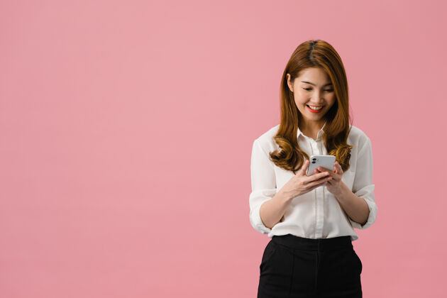 网络年轻的亚洲女士用手机 表情积极 笑容灿烂 穿着休闲服 感觉幸福 孤立地站在粉色背景上肖像女性使用