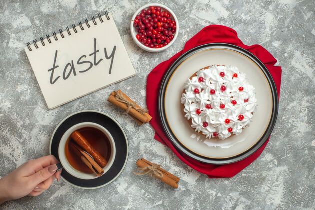 茶奶油蛋糕装饰着红色毛巾上的水果和一杯红茶与肉桂酸橙醋栗在一个白色的锅旁边的螺旋笔记本在冰的背景笔记本杯子醋栗