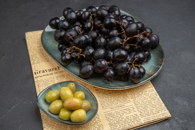 深色在黑暗的背景下 从一张旧报纸上俯瞰新鲜的有机绿橄榄和一捆捆黑葡萄有机橄榄食物