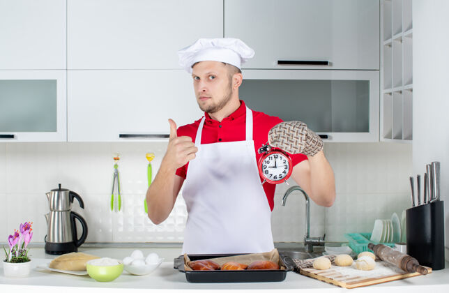 持有人年轻自信的男厨师的正面图戴着支架拿着钟 在白色厨房做着ok的手势男厨师成人微笑