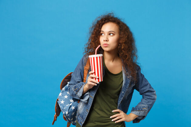 苏打水年轻的非洲裔美国女孩十几岁的学生在牛仔服装 背包举行纸杯隔离在蓝色的墙壁上人手塑料