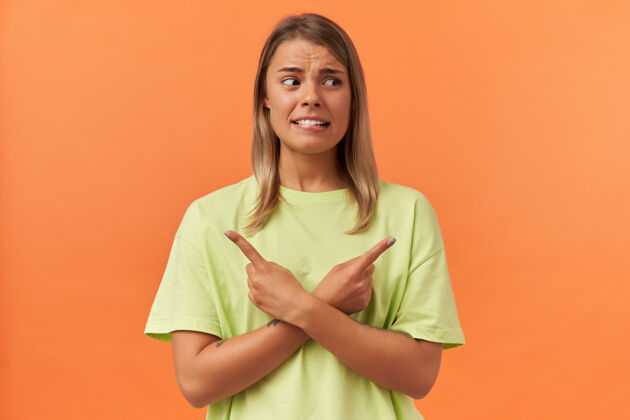指着一个穿着黄色t恤的困惑焦虑的年轻女人咬着嘴唇 用手指指着两边 隔着橙色的墙不开心T恤方向