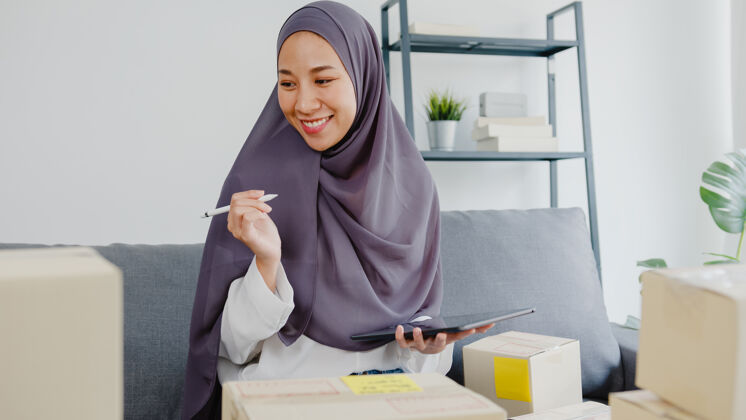 电子商务年轻的女商人检查产品采购订单的库存和保存到平板电脑工作在家办公印尼语卖家头巾