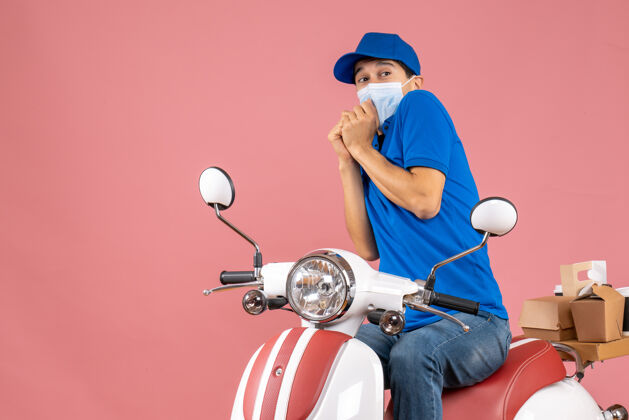 摩托车俯视图：戴着医用面罩戴着帽子的快递员坐在滑板车上 在柔和的桃色背景下传递订单坐命令面具