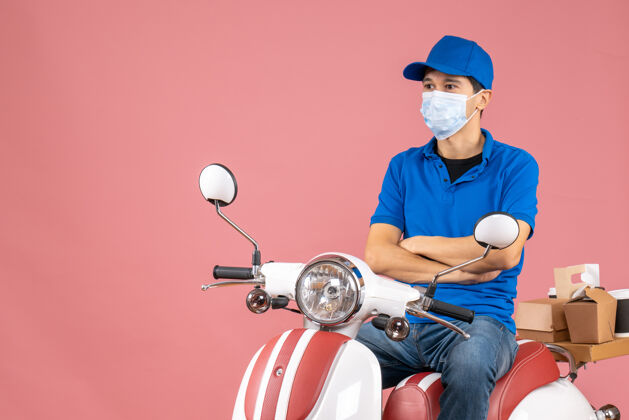 背景俯视图中的快递员戴着医疗面罩 戴着帽子 坐在滑板车上 在柔和的桃色背景下仔细地看着一些东西医疗面具摩托车