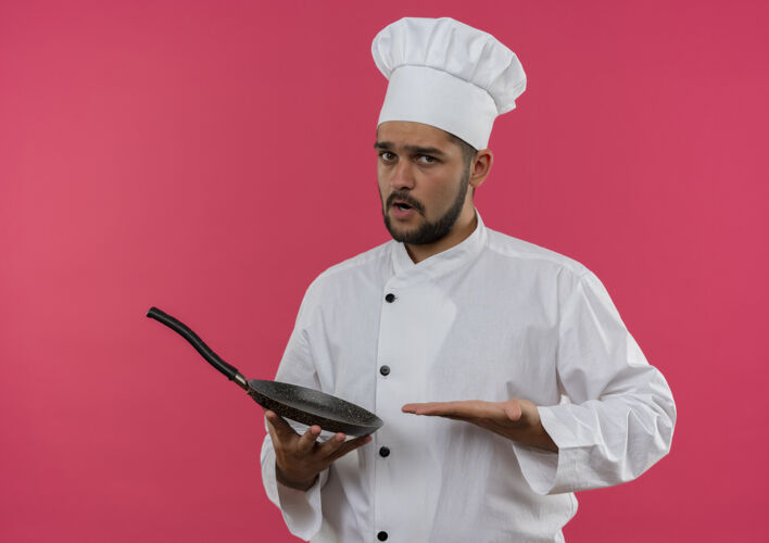 厨师穿着厨师制服的年轻男厨师手拿并指着隔离在粉红色墙上的煎锅男烹饪薯条