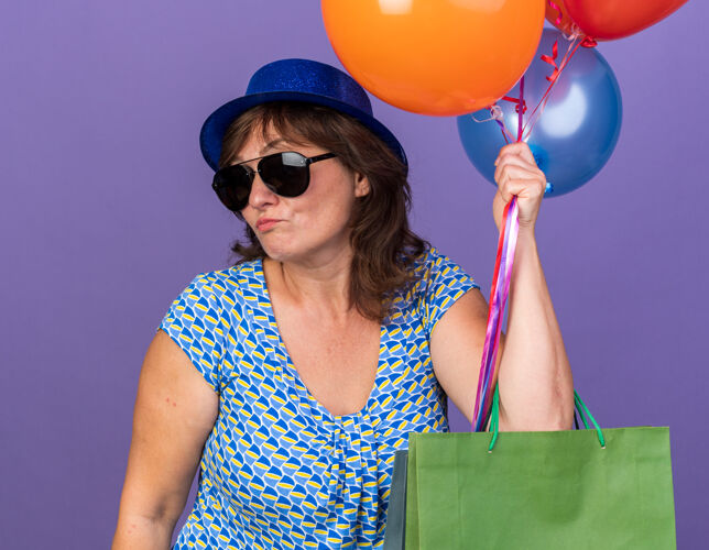 中间中年妇女戴着派对帽 戴着眼镜 手里拿着一堆五颜六色的气球和纸袋 手里拿着礼物 神情困惑不安地站在紫色的墙上庆祝生日派对站帽子年龄