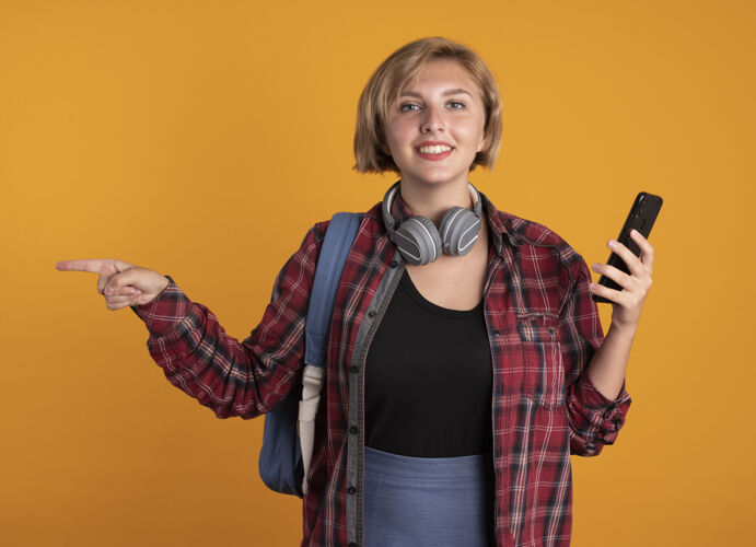 壁板微笑着的年轻斯拉夫女学生戴着耳机 背着背包 拿着电话 指着旁边电话穿空间