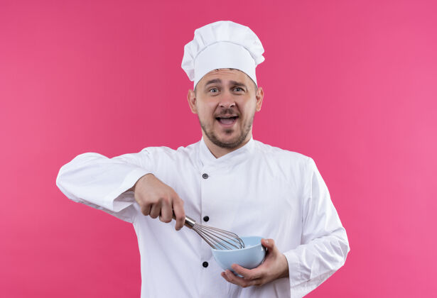 碗年轻帅气的厨师穿着厨师制服 手里拿着搅拌器和碗 孤立地站在粉红色的墙上烹饪年轻持有